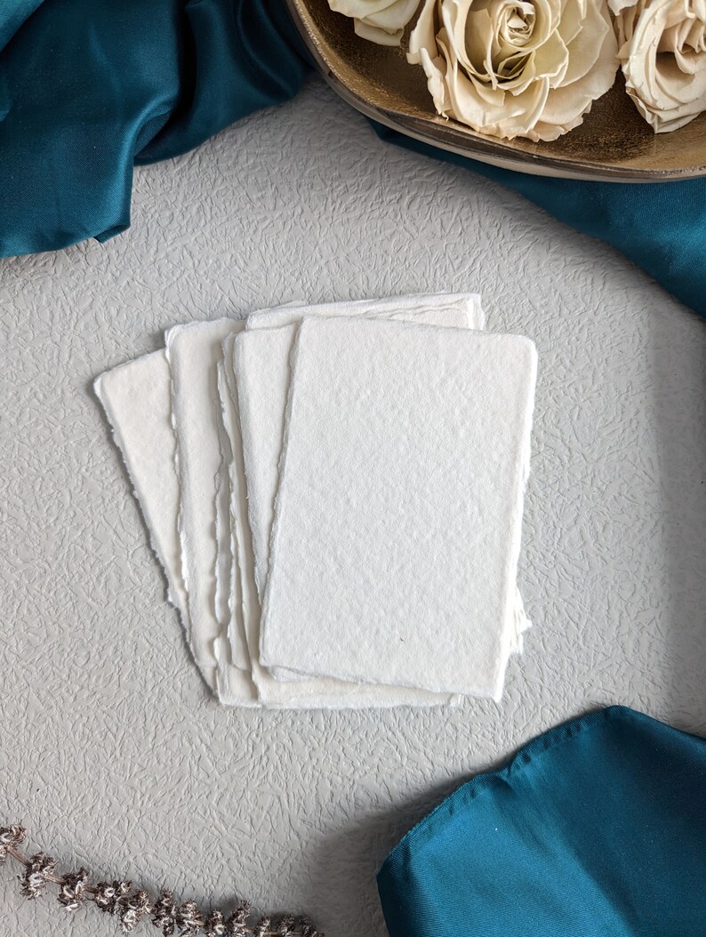 May include: A stack of five white, textured, handmade paper cards with deckled edges. The cards are on a light gray textured surface with a teal blue fabric in the background.