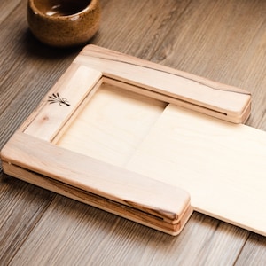 May include: A wooden tray with a sliding panel, perfect for serving snacks or holding small items. The tray is made of light wood with a natural finish and has a small logo on the side.