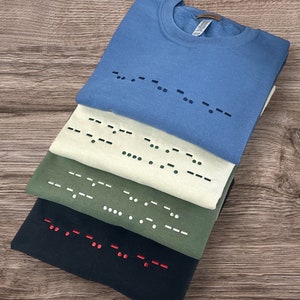 Peut inclure: Quatre sweat-shirts pliés de couleurs variées : bleu, crème, vert olive et noir. Chaque chemise présente un motif en code Morse sur le devant. Le code Morse est noir sur les chemises bleues et crème, blanc sur la chemise vert olive et rouge sur la chemise noire.