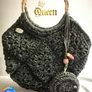 Puede incluir: Bolso de crochet gris hecho a mano con asa de bambú. El bolso presenta un patrón de puntada texturizada y un pequeño monedero a juego sujeto con una borla y cuentas. La palabra "Queen" está escrita en oro en el bolso.