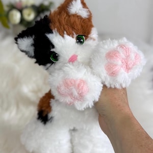 May include: A plush calico cat toy featuring white, brown, and black fur patches. The stuffed animal has bright green eyes, a small pink nose, and prominent white paws with textured pink paw pads. The fur appears soft and fluffy throughout.