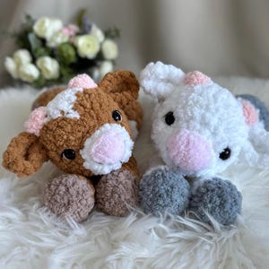Peut inclure: Deux veaux en peluche. L'un est marron et blanc, l'autre est blanc. Les deux ont des nez et des oreilles roses et des yeux foncés. Les veaux sont couchés sur une surface blanche et moelleuse. Le veau brun a des pattes marron et beige, et le veau blanc a des pattes grises.