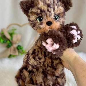May include: A plush toy animal with brown and tan mottled fur, large bright green eyes, and a small black nose. The creature features dark brown fur on its paws with light pink, rounded paw pads visible on the front-facing limbs.