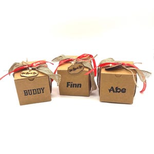 Pode incluir: Tr&ecirc;s caixas de presente quadradas de papel&atilde;o na cor marrom claro. Cada caixa apresenta um nome impresso em preto: BUDDY, Finn e Abe. As caixas s&atilde;o decoradas com uma fita de juta, uma fita vermelha fina e uma etiqueta de madeira Old Rural Life.
