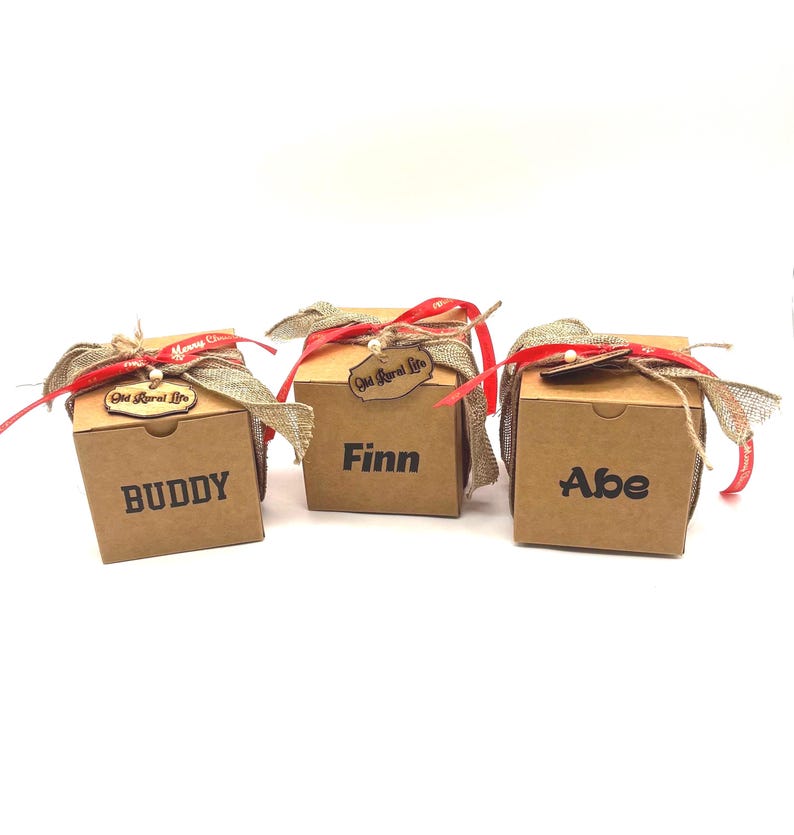 Pode incluir: Tr&ecirc;s caixas de presente quadradas de papel&atilde;o na cor marrom claro. Cada caixa apresenta um nome impresso em preto: BUDDY, Finn e Abe. As caixas s&atilde;o decoradas com uma fita de juta, uma fita vermelha fina e uma etiqueta de madeira Old Rural Life.