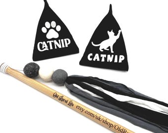 Könnte beinhalten: Katzenspielzeug. Schwarze dreieckige Katzenminze-Beutel mit weißen Pfoten- und Katzengrafiken und dem Wort "CATNIP". Ein Holzstab mit Filzkugeln und Stoffbändern in Grau, Schwarz und Weiß.
