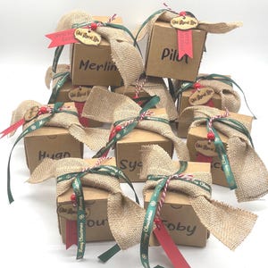 Pode incluir: Uma cole&ccedil;&atilde;o de caixas de presente quadradas de papel&atilde;o, cada uma com uma fita de juta, uma fita verde com o texto Merry Christmas, uma fita vermelha e uma etiqueta de madeira com a inscri&ccedil;&atilde;o Old Rural Life. Cada caixa tem um nome em preto.