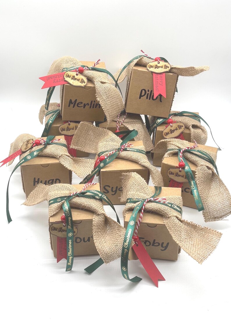 Pode incluir: Uma cole&ccedil;&atilde;o de caixas de presente quadradas de papel&atilde;o, cada uma com uma fita de juta, uma fita verde com o texto Merry Christmas, uma fita vermelha e uma etiqueta de madeira com a inscri&ccedil;&atilde;o Old Rural Life. Cada caixa tem um nome em preto.