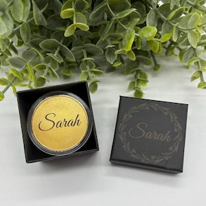 May include: A black gift box containing a gold-coloured compact with the name "Sarah" inscribed on it. A second black box, also inscribed with "Sarah", is visible. Green foliage is in the background.
