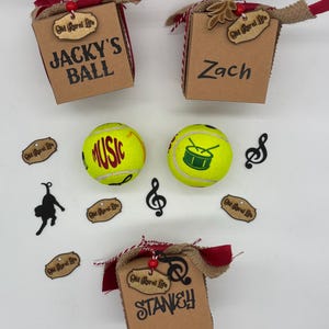 Pode incluir: Tr&ecirc;s caixas de presente de papel&atilde;o marrom com barbante vermelho e branco, fita de juta e etiquetas de madeira com a inscri&ccedil;&atilde;o Old Rural Life. As caixas s&atilde;o personalizadas com os nomes Jacky's Ball, Zach e Stanley. Duas bolas de t&ecirc;nis amarelas apresentam gr&aacute;ficos com temas musicais.