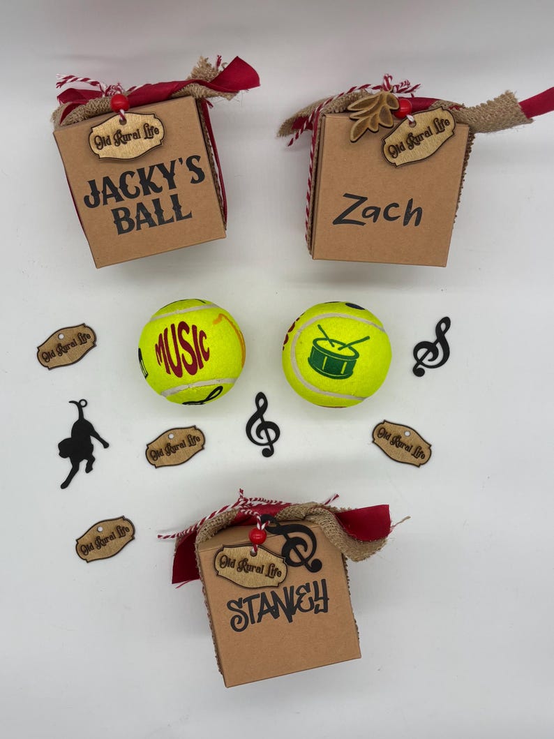 Pode incluir: Tr&ecirc;s caixas de presente de papel&atilde;o marrom com barbante vermelho e branco, fita de juta e etiquetas de madeira com a inscri&ccedil;&atilde;o Old Rural Life. As caixas s&atilde;o personalizadas com os nomes Jacky's Ball, Zach e Stanley. Duas bolas de t&ecirc;nis amarelas apresentam gr&aacute;ficos com temas musicais.