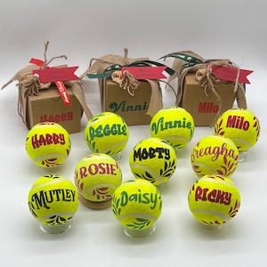 Pode incluir: Dez bolas de t&eacute;nis amarelo-brilhante, cada uma personalizada com um nome em tinta vermelha ou verde e padr&otilde;es de folhas decorativas, dispostas &agrave; frente de tr&ecirc;s pequenas caixas de presente de cart&atilde;o castanho com fitas de serapilheira e etiquetas.