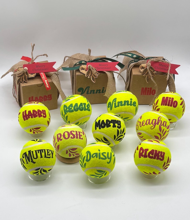Pode incluir: Dez bolas de t&eacute;nis amarelo-brilhante, cada uma personalizada com um nome em tinta vermelha ou verde e padr&otilde;es de folhas decorativas, dispostas &agrave; frente de tr&ecirc;s pequenas caixas de presente de cart&atilde;o castanho com fitas de serapilheira e etiquetas.