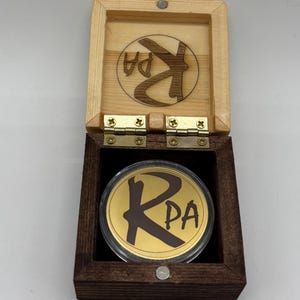 May include: A square wooden box with a dark brown base and a light natural wood lid, featuring a gold-coloured circular coin inside a clear protective case. The lid interior and the coin both display a dark brown logo with the letters KPA.