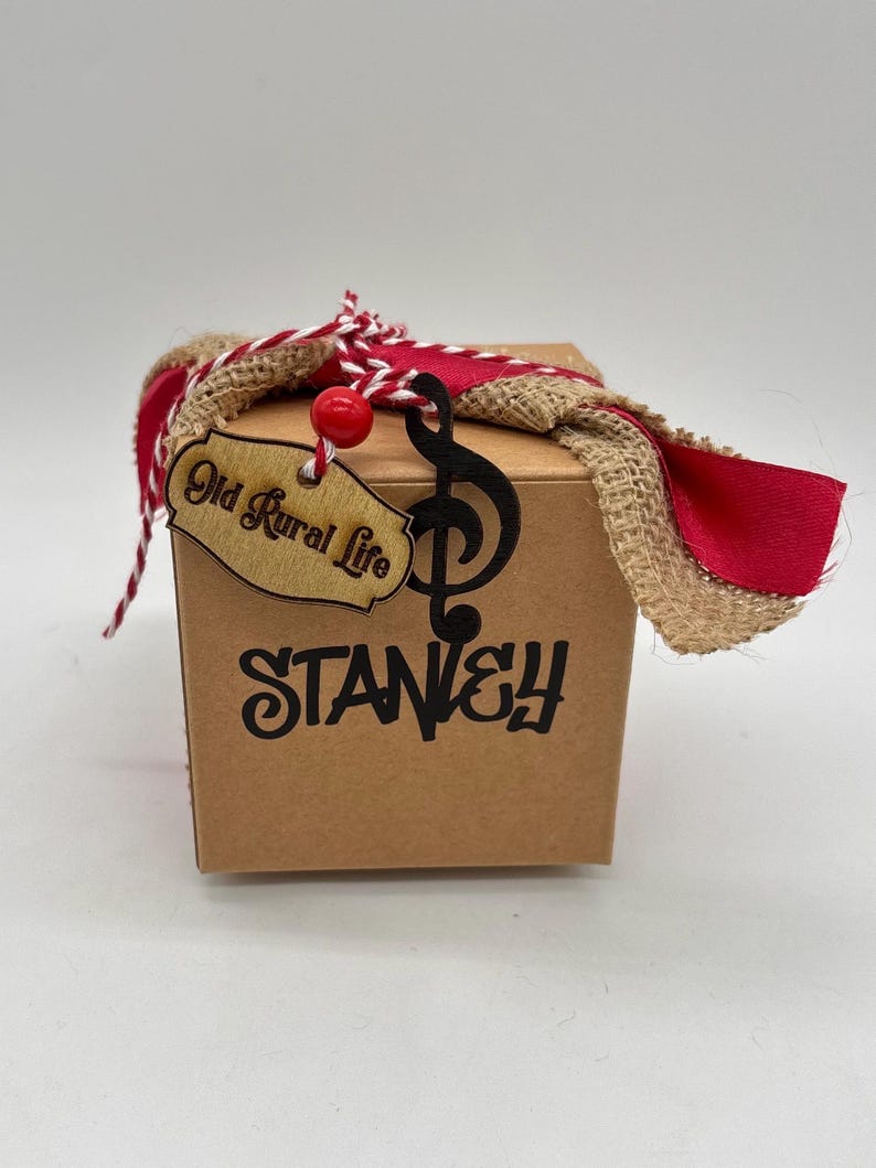 Pode incluir: Uma caixa quadrada de papel&atilde;o com o texto STANLEY impresso em preto. Um recorte de clave de sol preta e uma etiqueta de madeira com a inscri&ccedil;&atilde;o Old Rural Life est&atilde;o presos com barbante vermelho e branco, fita vermelha e tecido de juta.