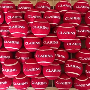 Pode incluir: Uma cole&ccedil;&atilde;o de bolas de t&eacute;nis vermelhas dispostas numa grelha, cada uma com o texto branco CLARINS impresso no centro. As bolas t&ecirc;m uma textura felpuda e linhas curvas brancas t&iacute;picas da constru&ccedil;&atilde;o das bolas de t&eacute;nis, preenchendo o quadro.