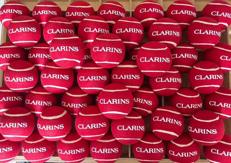 Pode incluir: Uma cole&ccedil;&atilde;o de bolas de t&eacute;nis vermelhas dispostas numa grelha, cada uma com o texto branco CLARINS impresso no centro. As bolas t&ecirc;m uma textura felpuda e linhas curvas brancas t&iacute;picas da constru&ccedil;&atilde;o das bolas de t&eacute;nis, preenchendo o quadro.