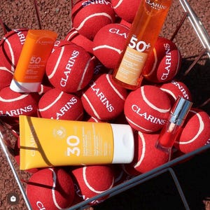 Pode incluir: Uma cesta de arame cheia de bolas de t&eacute;nis vermelhas com o log&oacute;tipo Clarins, contendo um spray Clarins SPF 50+, um stick Clarins SPF 50, um tubo Clarins SPF 30 e um pequeno frasco de &oacute;leo labial vermelho com tampa prateada.