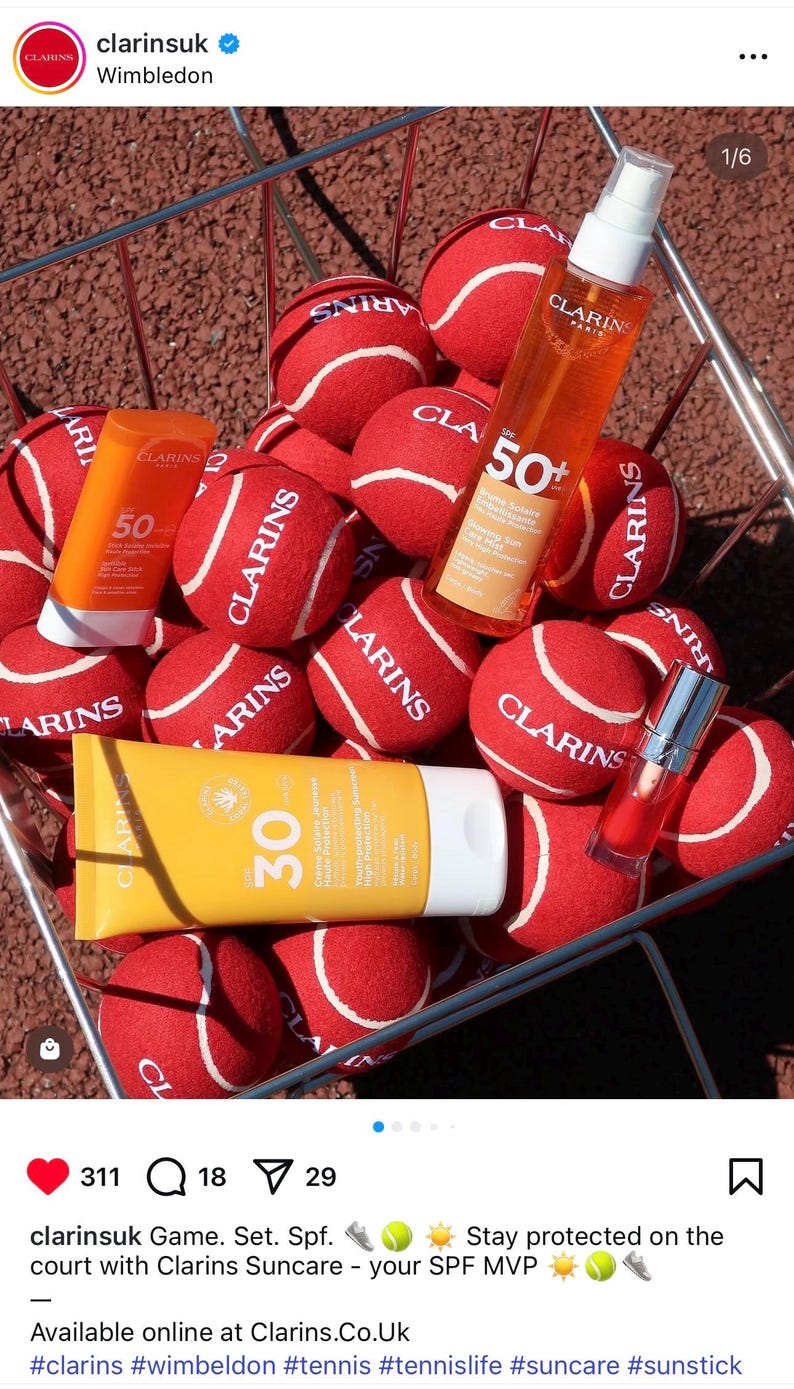 Pode incluir: Uma cesta de arame cheia de bolas de t&eacute;nis vermelhas com o log&oacute;tipo Clarins, contendo um spray Clarins SPF 50+, um stick Clarins SPF 50, um tubo Clarins SPF 30 e um pequeno frasco de &oacute;leo labial vermelho com tampa prateada.