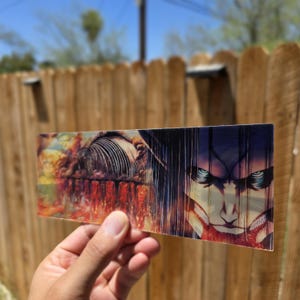 May include: A rectangular, transparent acrylic print featuring a vibrant anime-style illustration. The artwork depicts a dramatic scene with a character's face and a structure with a red and orange background. The print is held against a wooden fence.