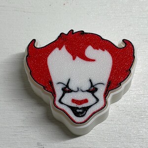 May include: A white plastic cookie cutter shaped like the head of a clown with a textured red hair design, a white face, black outlined eyes, and a black and red mouth. The object has a raised edge and a flat base for cutting dough shapes.