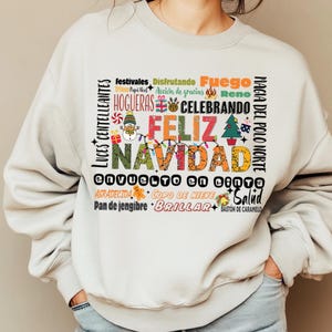 May include: A white sweatshirt with a festive Christmas design featuring the words "Feliz Navidad" in colorful letters surrounded by other holiday-themed words in Spanish. The design includes images of a snowman, a Christmas tree, a candy cane, and a gingerbread man.