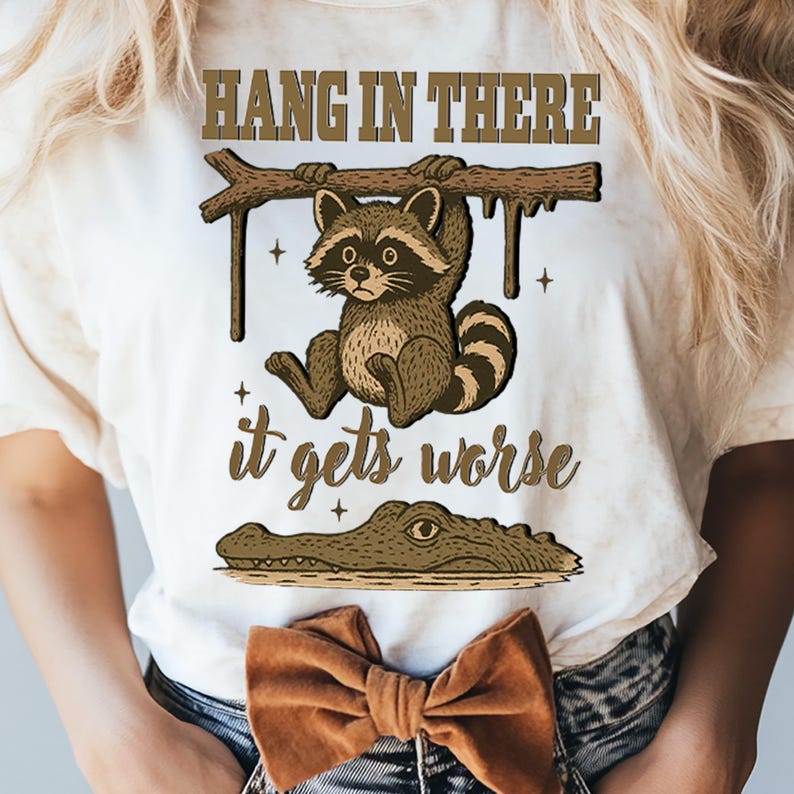 以下が含まれることがあります： オフホワイトのTシャツ。木にぶら下がっているアライグマのイラストがあり、上部に「HANG IN THERE」、下部に「it gets worse」と書かれています。ワニが水中に描かれています。茶色のリボンが見えます。