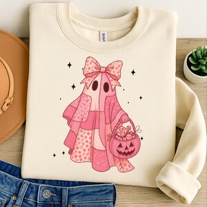 以下が含まれることがあります： クリーム色のスウェットシャツ。ピンクのパッチワークデザインのゴーストキャラクターが特徴です。ゴーストは大きなリボンをつけ、キャンディーの入ったジャック・オー・ランタンのバスケットを持っています。黒い星がゴーストを囲み、お祭り気分なハロウィーンのテーマを作り出しています。