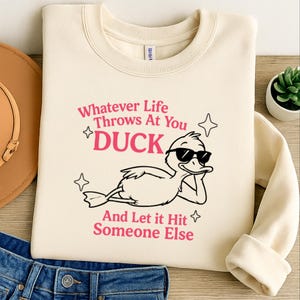 May include: Cream-colored sweatshirt with the text "Whatever Life Throws At You DUCK And Let it Hit Someone Else" in pink. A cartoon duck wearing sunglasses is featured in the design.