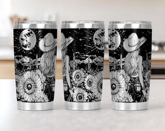 May include: Three stainless steel travel tumblers with a black and white illustration. The design features a woman in a cowboy hat, sunflowers, a full moon, and a desert landscape. The tumblers have a silver rim and lid.