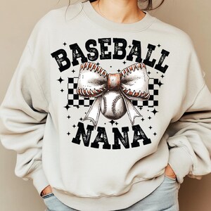 Op de afbeelding: Een wit sweatshirt met de tekst "Baseball Nana" in zwarte letters. Een baseball met een witte strik is gecentreerd op het sweatshirt.