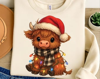 May include: Cream-colored sweatshirt featuring a cartoon Highland cow wearing a Santa hat, a black and white plaid scarf, and a string of colorful Christmas lights. The cow has brown fur and large, expressive eyes. A festive holiday design.