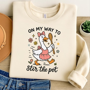 Puede incluir: Sudadera color crema con un dibujo de un ganso de dibujos animados con un delantal rosa y una cuchara de madera. El texto "ON MY WAY TO" y "stir the pot" están encima y debajo del ganso, con flores y estrellas coloridas.