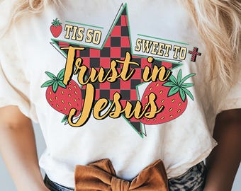 May include: Cream-colored t-shirt featuring a star-shaped graphic with a red and black checkered pattern. The graphic includes the text "Trust in Jesus" in yellow, along with strawberries and the phrases "Tis So" and "Sweet To".