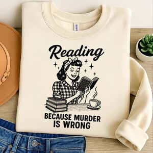 May include: Cream-colored sweatshirt with a graphic of a woman reading a book, a stack of books, and a coffee cup. The text reads "Reading BECAUSE MURDER IS WRONG" in black script.