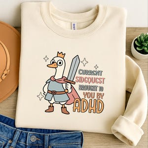 Op de afbeelding: Crèmekleurige sweatshirt met een cartoon gans met een kroon, cape en zwaard. De tekst luidt "Current Sidequest Brought to You by ADHD". Het ontwerp bevat sterren.