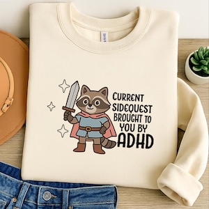 Op de afbeelding: Crèmekleurige sweatshirt met een cartoon wasbeer als ridder, die een zwaard vasthoudt. De tekst luidt "Current Sidequest Brought to You by ADHD". Het ontwerp bevat sterren en een roze cape.