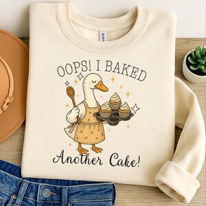 May include: Cream-colored sweatshirt featuring a cartoon duck wearing an apron and holding a wooden spoon and a tray of cupcakes. The text reads "Oops! I Baked Another Cake!" in black script. The design includes star accents.