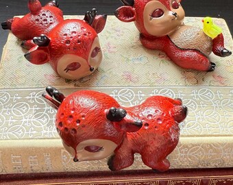 May include: Three red ceramic deer figurines with white eyes and black pupils. The deer are in various poses, including lying down and sitting up. One deer is holding a small yellow bird figurine.