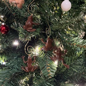 May include: Brown frog-shaped Christmas ornaments hanging on a green Christmas tree. The ornaments have gold-colored wire hangers. A red and white ornament and a white ball ornament are also on the tree.