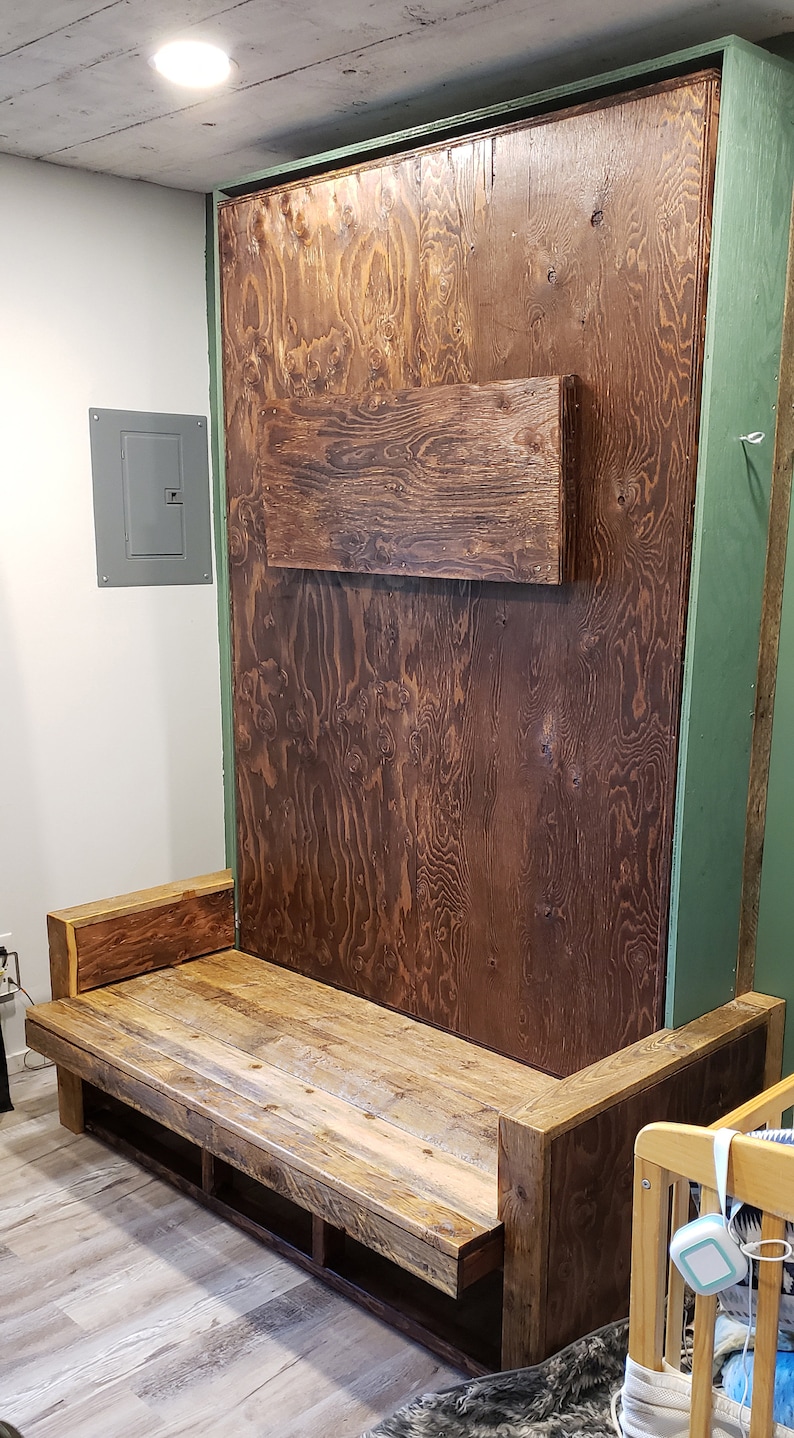 May include: A wooden bench with a built-in storage shelf. The bench is made of reclaimed wood and has a rustic look. The bench is in front of a wall with a wooden panel that is hinged to fold down, revealing a bed.