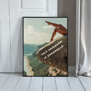 Peut inclure: Impression d'art encadr&eacute;e avec un paysage. On voit les jambes d'une personne sur un rocher. Le texte "THIS DAYDREAM IS DANGEROUS" est en blanc. L'impression est contre un mur blanc.