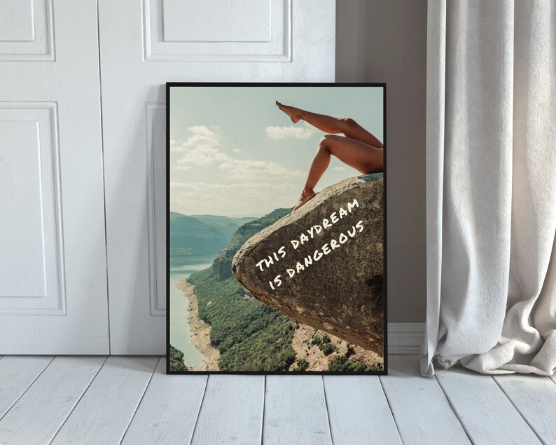 Peut inclure: Impression d'art encadr&eacute;e avec un paysage. On voit les jambes d'une personne sur un rocher. Le texte "THIS DAYDREAM IS DANGEROUS" est en blanc. L'impression est contre un mur blanc.