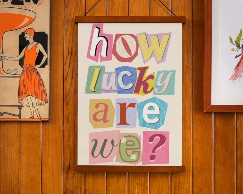 Op de afbeelding: Een ingelijst print met de woorden "how lucky are we?" in kleurrijke, uitgesneden letters op een witte achtergrond.