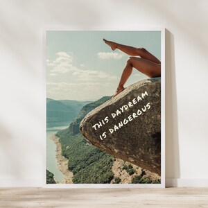 Peut inclure: Une affiche encadr&eacute;e repr&eacute;sentant un paysage avec un lac et des montagnes. L'image comprend les jambes d'une personne sur un rocher avec le texte "THIS DAYDREAM IS DANGEROUS" en blanc.