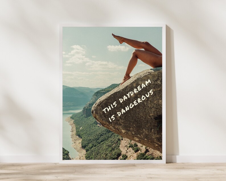 Peut inclure: Une affiche encadr&eacute;e repr&eacute;sentant un paysage avec un lac et des montagnes. L'image comprend les jambes d'une personne sur un rocher avec le texte "THIS DAYDREAM IS DANGEROUS" en blanc.