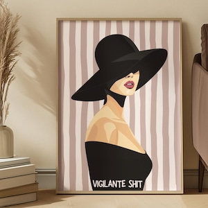 Può includere: Una stampa artistica incorniciata raffigurante una donna con cappello e abito neri, con la scritta "VIGILANTE SHIT" in basso. Lo sfondo presenta strisce verticali nei toni del rosa e del beige. L'opera d'arte è esposta in una stanza con un vaso di fiori secchi e una pila di libri.