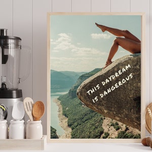 Peut inclure: Une affiche encadr&eacute;e repr&eacute;sentant un paysage avec un lac et des montagnes. Les jambes d'une personne sont visibles, s'&eacute;tendant vers le haut depuis un rocher. Le texte "THIS DAYDREAM IS DANGEROUS" est &eacute;crit en blanc sur le rocher.