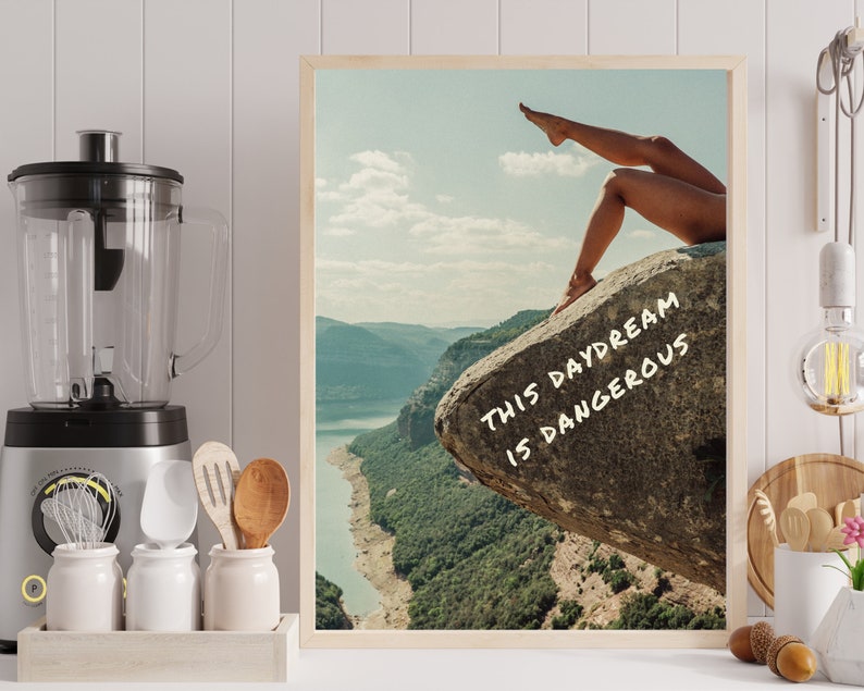 Peut inclure: Une affiche encadr&eacute;e repr&eacute;sentant un paysage avec un lac et des montagnes. Les jambes d'une personne sont visibles, s'&eacute;tendant vers le haut depuis un rocher. Le texte "THIS DAYDREAM IS DANGEROUS" est &eacute;crit en blanc sur le rocher.