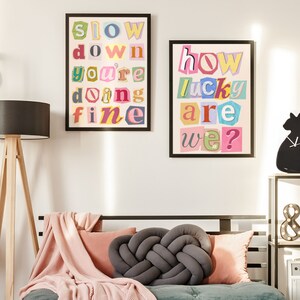 Op de afbeelding: Twee ingelijste prints met kleurrijke uitgesneden letters die "slow down you're doing fine" en "how lucky are we?" spellen, hangen aan een witte muur boven een grijze bank met een roze deken en een grijs geknoopt kussen.